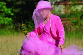 Ein Foto von Rosa von Praunheim in einem pinken Kleid mit pinkem Hut, von dem aus ein Schleier vor das Gesicht fällt. Rosa von Praunheim steht auf einer grünen Wiese. 